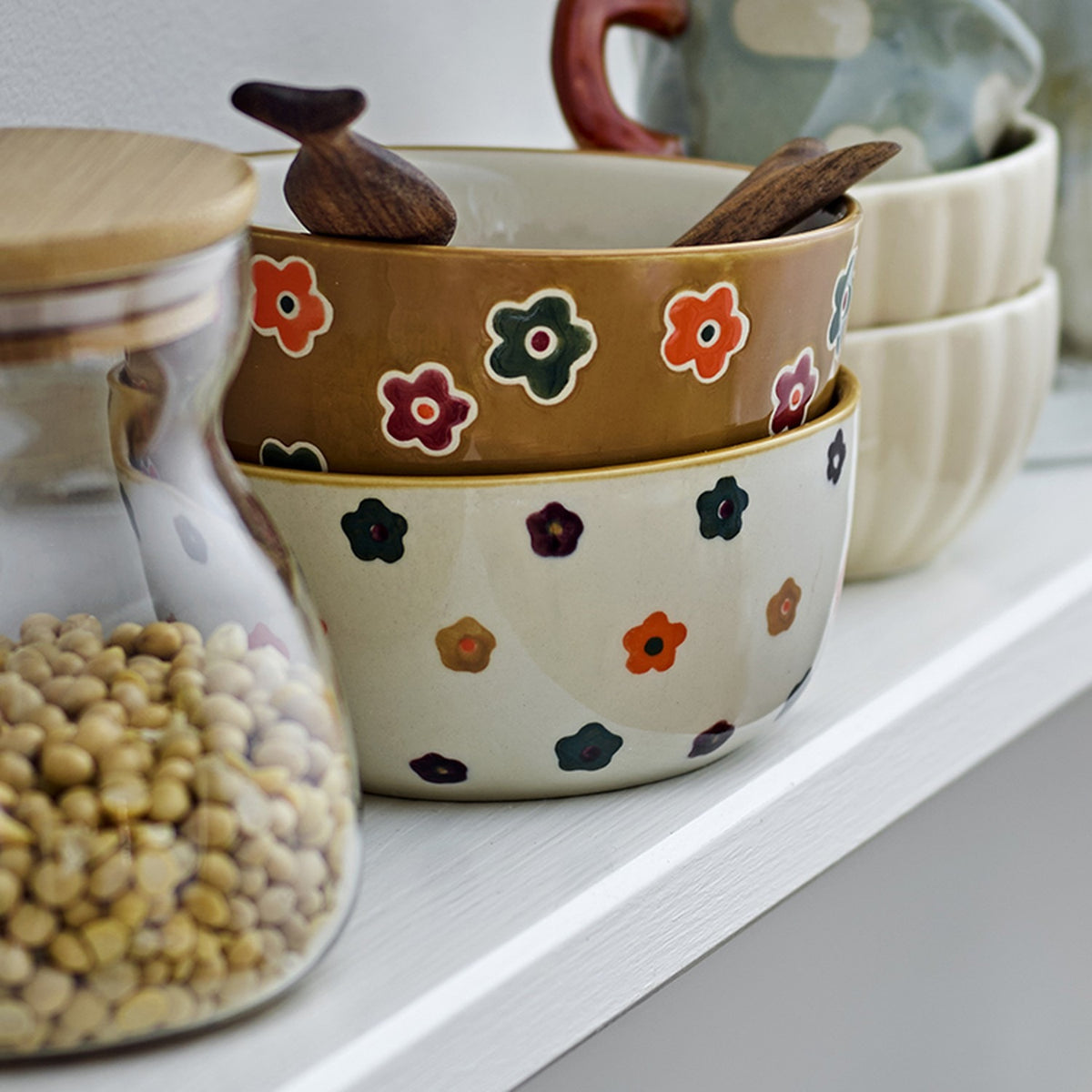 Addy Stoneware Bowl 12cm in cream, hand-decorated with small multicoloured flowers, stacked with other patterned bowls and wooden spoons on a shelf.