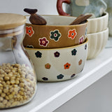 Addy Stoneware Bowl 12cm in cream, hand-decorated with small multicoloured flowers, stacked with other patterned bowls and wooden spoons on a shelf.