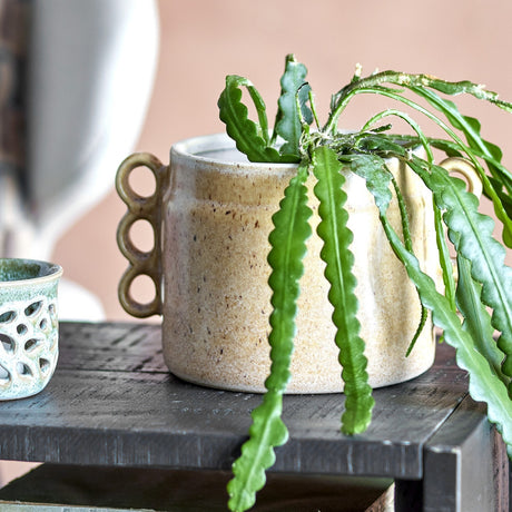 Souri Stoneware Plant Pot in beige, a round speckled ceramic planter with triple-loop side handles, shown on a dark wooden shelf with trailing green foliage.