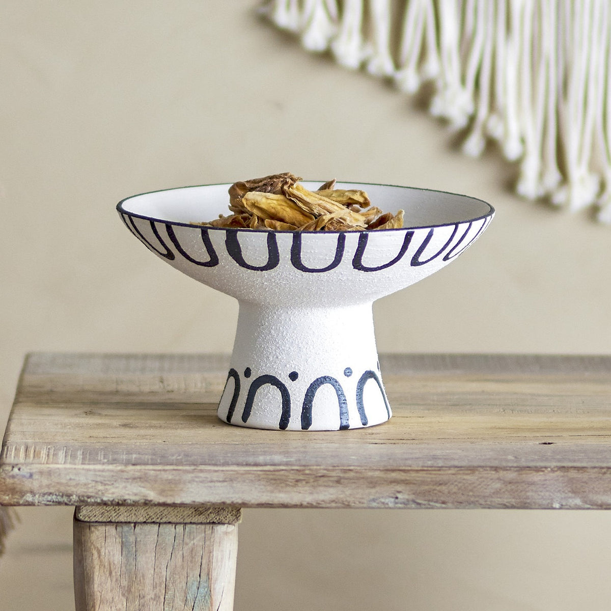 Mayotte Stoneware Pedestal Bowl 26cm in cream with navy loop and arch pattern, shown filled with dried flowers on a wooden bench.