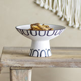 Mayotte Stoneware Pedestal Bowl 26cm in cream with navy loop and arch pattern, shown filled with dried flowers on a wooden bench.
