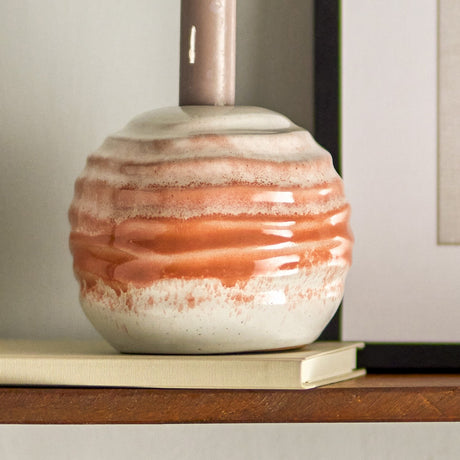 Paula Round Stoneware Candle Holder in orange, a squat ridged ceramic base with reactive glaze bands, shown holding a candle on a book beside a shelf.