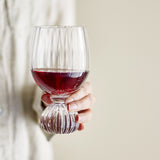 Hand holding the Taurin Round Wine Glass in clear ribbed glass with a short round foot, filled partway with red wine against a neutral background.