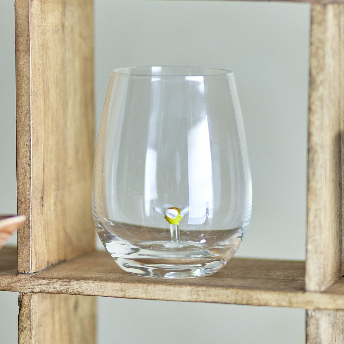 Misa Floral Tumbler in clear mouthblown glass with a stemless, rounded body and a small flower detail rising from the base, displayed on a wooden shelf.