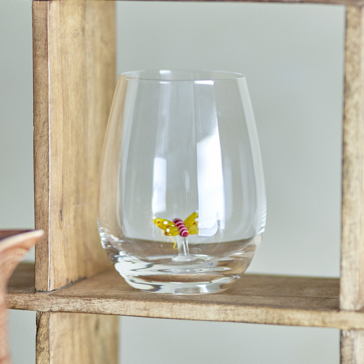 Misa Butterfly Tumbler in clear glass with a small yellow and pink glass butterfly raised on a stalk from the base, shown on a wooden shelf.