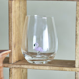 Misa Flamingo Tumbler in clear glass with a small pink flamingo standing on a stem inside, stemless rounded body on a wooden shelf.