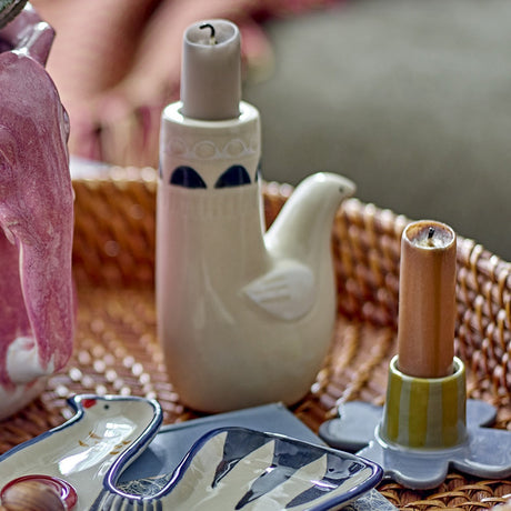 Trudy Bird Stoneware Candle Holder in cream ceramic, shaped like a bird with a tall taper candle column, shown on a woven tray among other decor pieces.