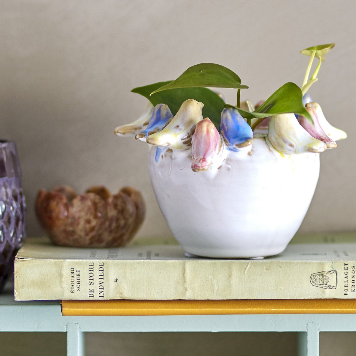 Eanna Stoneware Plant Pot in Multi, a round white ceramic planter with hand-painted birds around the rim, holding a leafy plant on a shelf with books and ornaments.