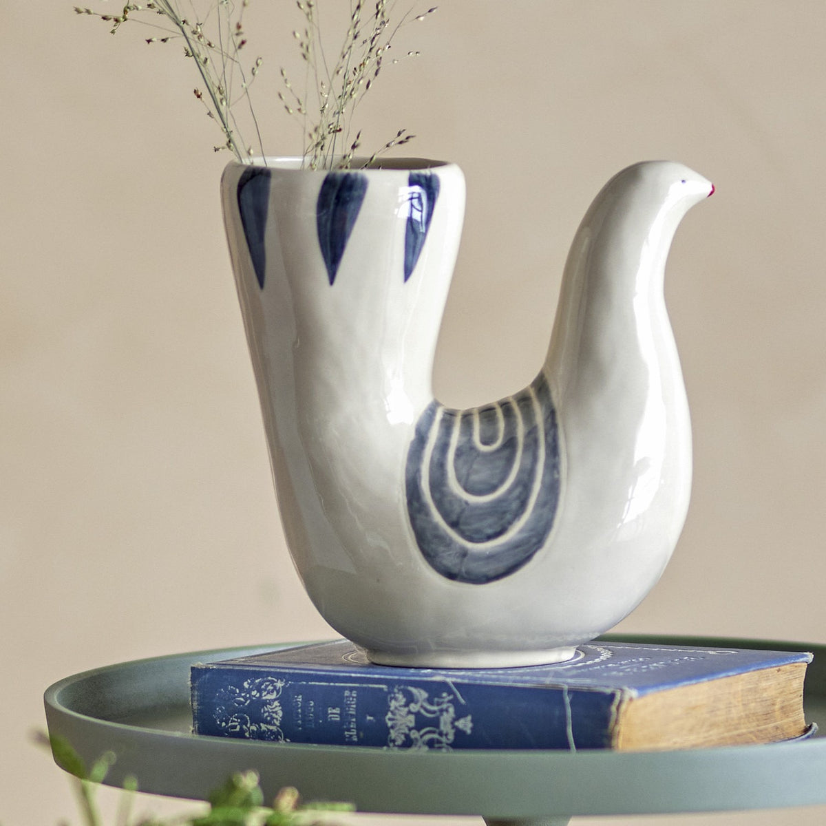 Trudy Bird Stoneware Vase in cream, shaped like a bird with a slender head and tall open tail holding dried stems, hand-painted blue wing and feather details, displayed on a book.