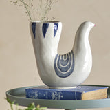 Trudy Bird Stoneware Vase in cream, shaped like a bird with a slender head and tall open tail holding dried stems, hand-painted blue wing and feather details, displayed on a book.