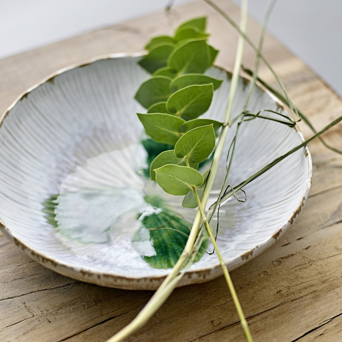 Mati Fluted Stoneware Bowl 29cm in white, a shallow ceramic serving bowl with radiating fluted texture, shown on a wooden surface with trailing leaves and grass stems inside.