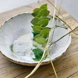 Mati Fluted Stoneware Bowl 29cm in white, a shallow ceramic serving bowl with radiating fluted texture, shown on a wooden surface with trailing leaves and grass stems inside.