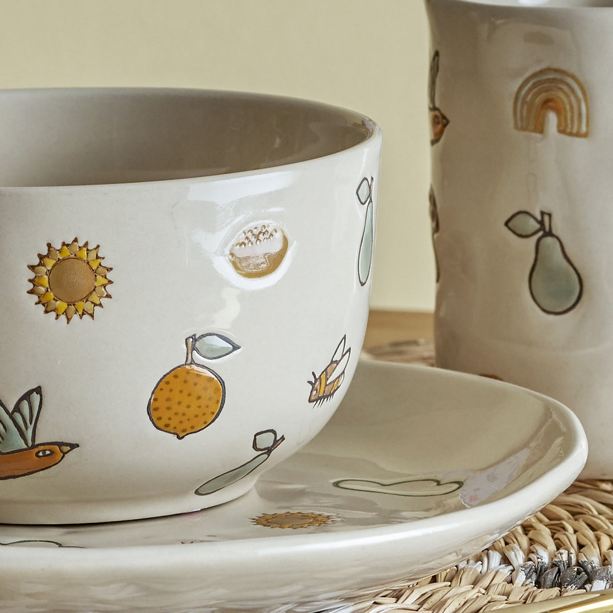 Agnes Stoneware Kids Dinner Set of 3 in cream, close-up of ceramic bowl, cup and plate hand-painted with sun, bird, pear and bee motifs.