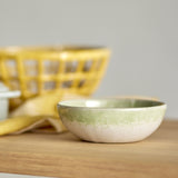 Paula Stoneware Bowl 16cm in cream, shallow ceramic bowl with a reactive green-toned glaze band below the rim, shown on a wooden surface with a yellow basket blurred in the background.
