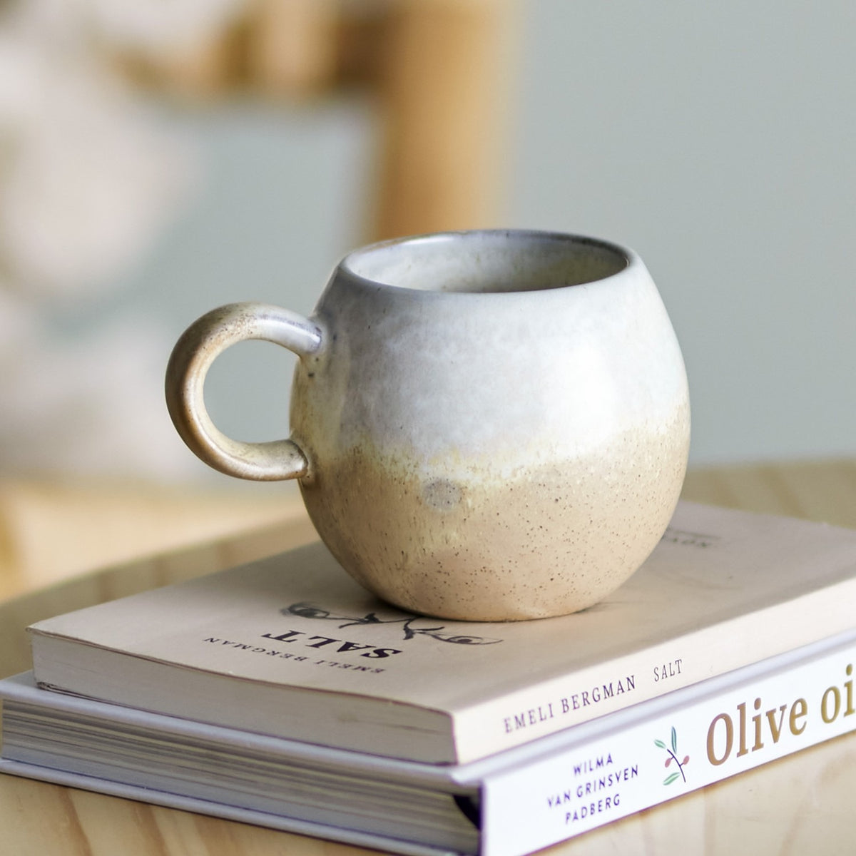 Paula Stoneware breakfast cup with loop handle in beige reactive glaze, rounded stoneware body, shown on stacked books in a kitchen setting.