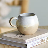 Paula Stoneware breakfast cup with loop handle in beige reactive glaze, rounded stoneware body, shown on stacked books in a kitchen setting.