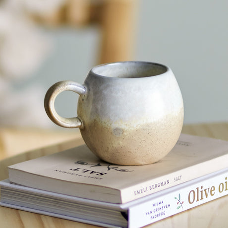 Paula Stoneware breakfast cup with loop handle in beige reactive glaze, rounded stoneware body, shown on stacked books in a kitchen setting.