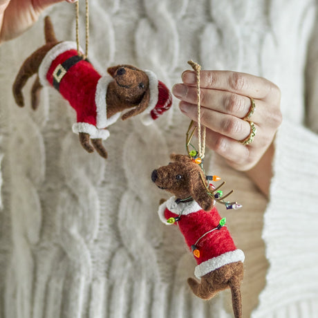 Set of two felted wool sausage dog Christmas ornaments in red Santa suits with fluffy trim, one with hat and belt, the other with antlers and fairy lights.