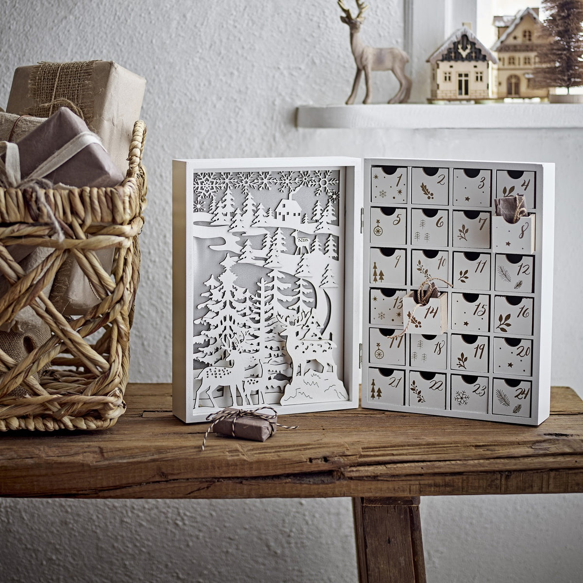 Elimar Wooden Advent Calendar in white, opened book-style with cut-out forest and stag scene on one side and 24 numbered drawers on the other, shown on a wooden bench.