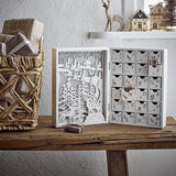 Elimar Wooden Advent Calendar in white, opened book-style with cut-out forest and stag scene on one side and 24 numbered drawers on the other, shown on a wooden bench.