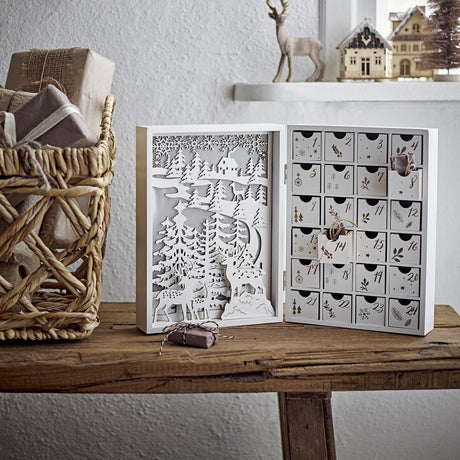 Elimar Wooden Advent Calendar in white, opened book-style with cut-out forest and stag scene on one side and 24 numbered drawers on the other, shown on a wooden bench.