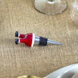 Lynn Glass Christmas Wine Stopper in red, a stainless steel cone with ribbed silicone rings topped by glass Father Christmas legs, shown beside a wine glass on a woven tablecloth.