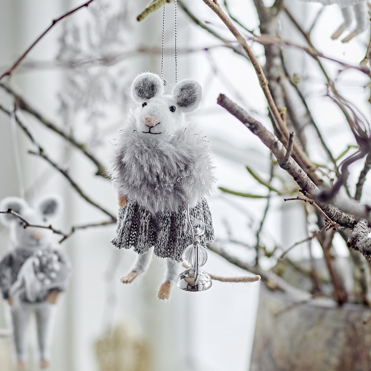 Peo Wool Christmas Tree Decoration mouse figure in grey, with a fluffy stole and knitted skirt, holding a small bell, hanging among bare branches.