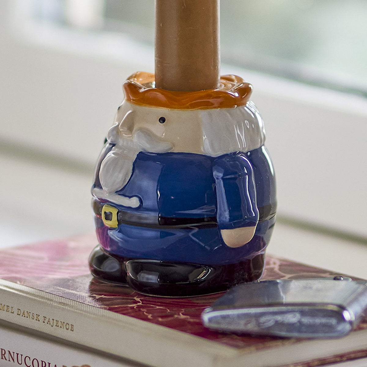 Feast Nutcracker stoneware Christmas candle holder in multicoloured ceramic, shaped like a nutcracker with blue coat and belt, holding a taper candle, shown on a book.
