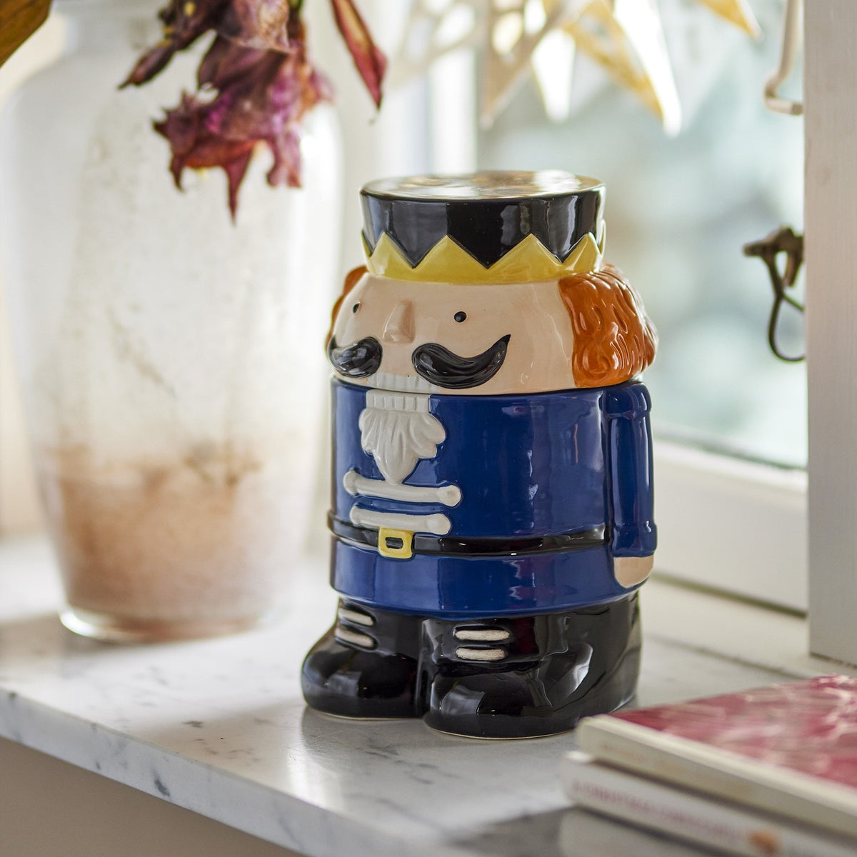 Feast Nutcracker Christmas storage jar with lid, 900ml ceramic design in navy, black and tan with crown and beard detail, shown on a marble shelf.
