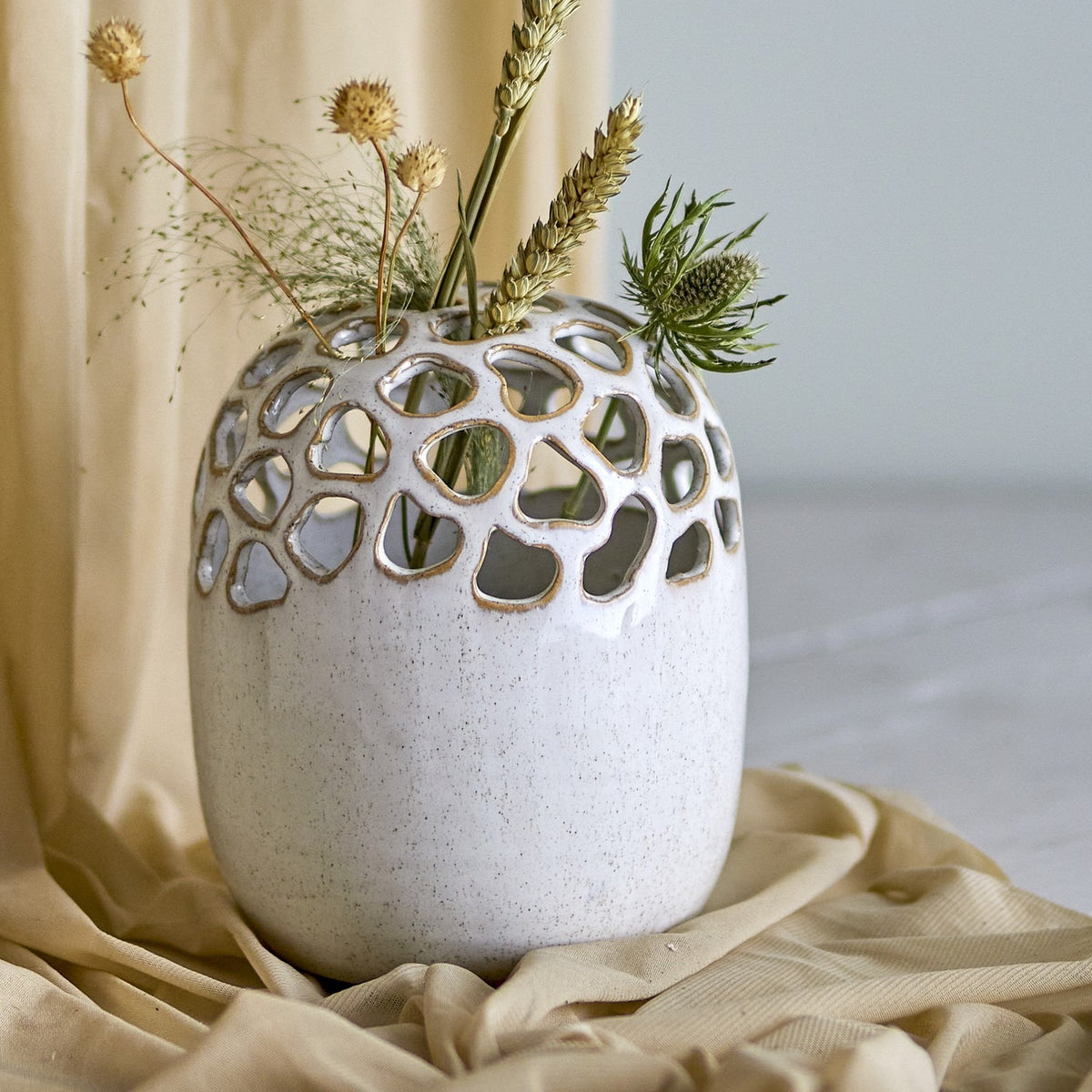 Elissa Speckled Stoneware Vase in cream, rounded body with pebble-shaped cut-out openings around the domed top, shown holding dried stems and foliage against a neutral backdrop.