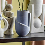 Norbert Stoneware Vase 18.5cm in mottled blue with a diagonal raised band and vertical ridge, holding leafy stems, displayed on a shelf among stone and ceramic ornaments.