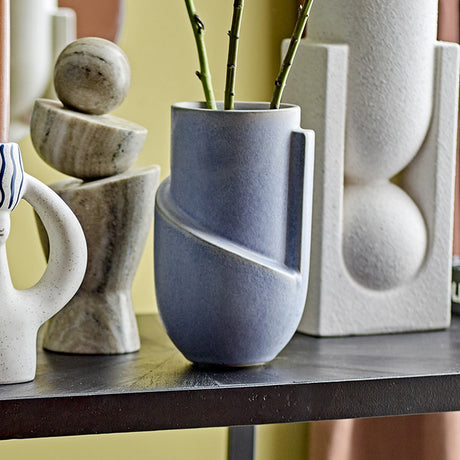 Norbert Stoneware Vase 18.5cm in mottled blue with a diagonal raised band and vertical ridge, holding leafy stems, displayed on a shelf among stone and ceramic ornaments.