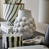 Poldor Stoneware Vase 19cm in cream with a raised bobble-textured middle and plain neck, shown on a dark table with striped boxes and a planter in the background.