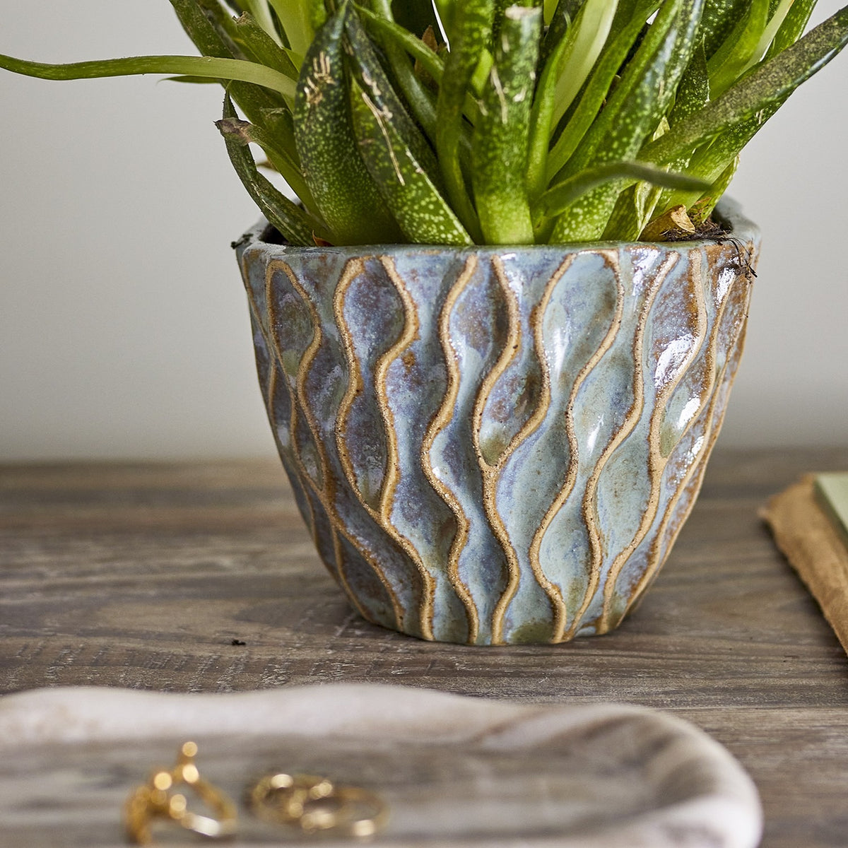 Feren Stoneware Plant Pot in blue, a small ceramic planter with wavy raised texture and speckled glaze, shown holding a green plant on a wooden surface.
