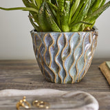 Feren Stoneware Plant Pot in blue, a small ceramic planter with wavy raised texture and speckled glaze, shown holding a green plant on a wooden surface.