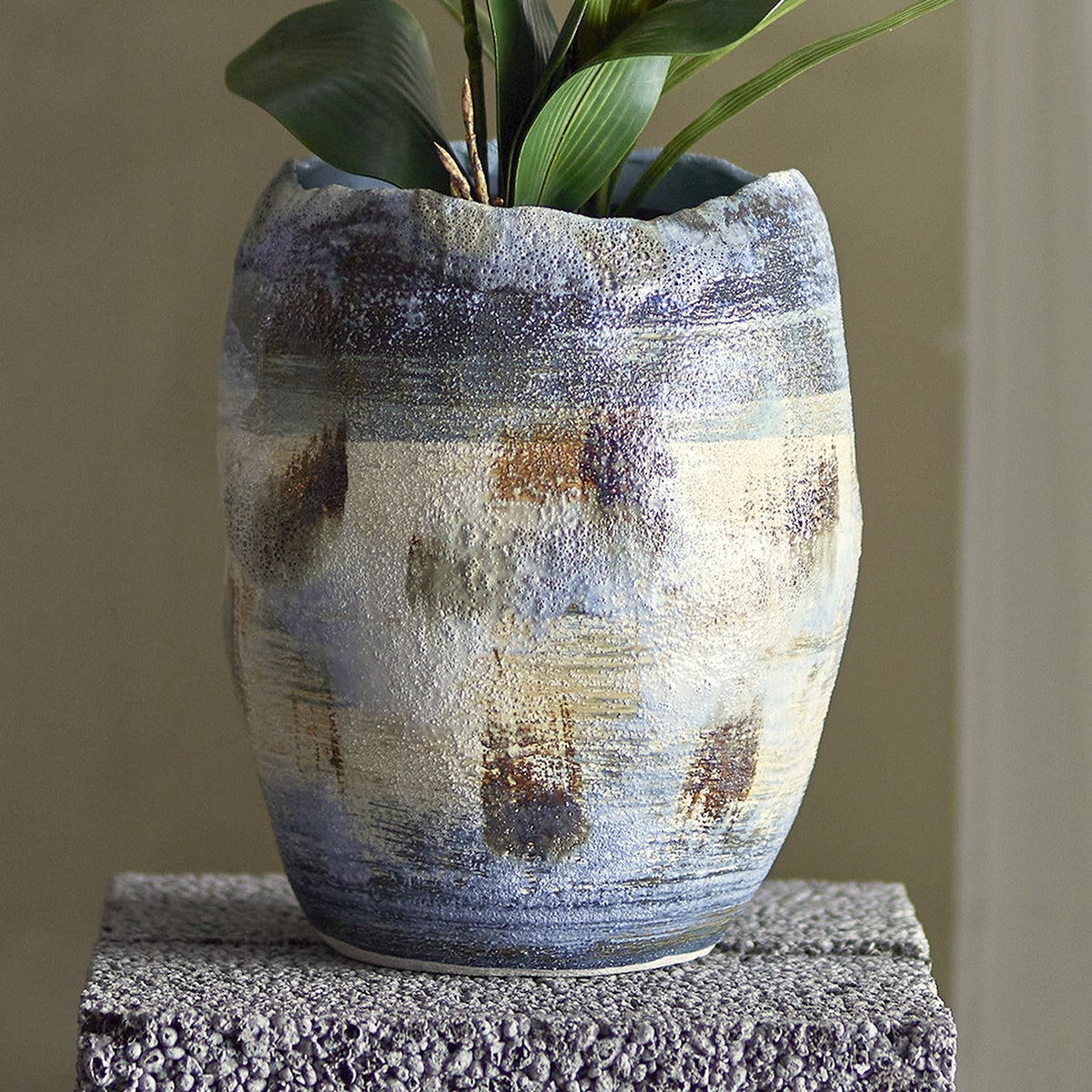 Nienna Stoneware Vase 31cm in Blue, a rustic ceramic vase with an uneven wavy rim and speckled blue glaze in bands, shown holding green foliage on a textured grey plinth.