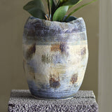 Nienna Stoneware Vase 31cm in Blue, a rustic ceramic vase with an uneven wavy rim and speckled blue glaze in bands, shown holding green foliage on a textured grey plinth.