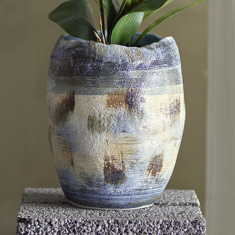 Nienna Stoneware Vase 31cm in Blue, a rustic ceramic vase with an uneven wavy rim and speckled blue glaze in bands, shown holding green foliage on a textured grey plinth.