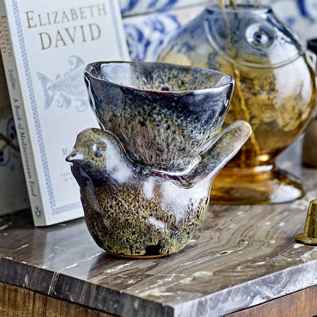 Brown stoneware bird ornament with raised tail holding a wide-rimmed bowl, glossy speckled glaze, displayed on a marble shelf with a book and amber glass vase.
