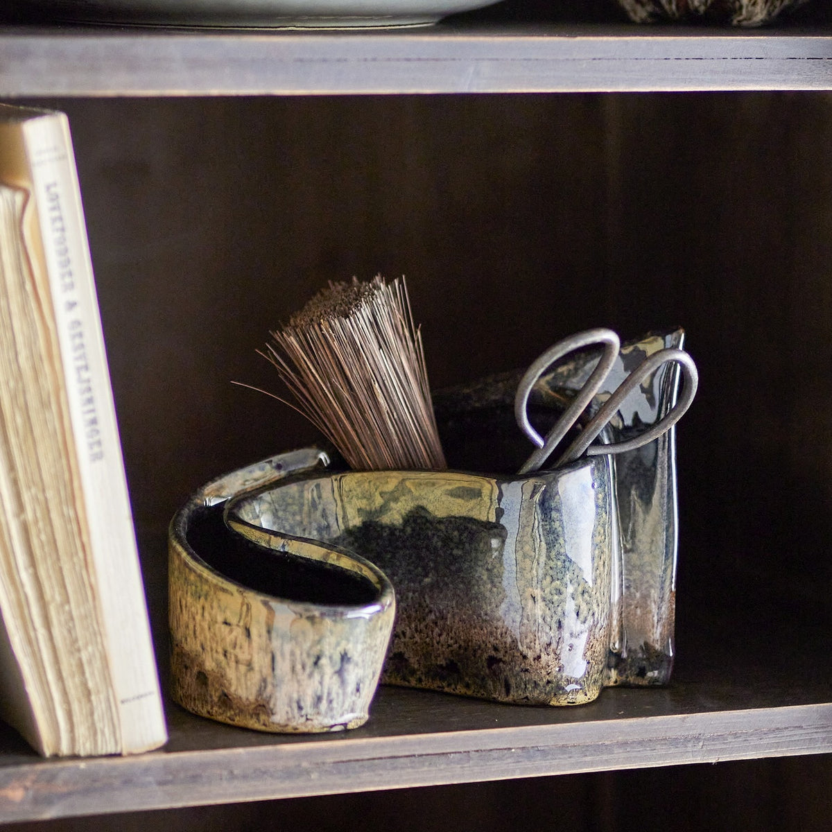 Phil Stoneware Ornament in brown, a curled ceramic piece with open hollows, shown on a shelf holding scissors and dried stems beside a book.