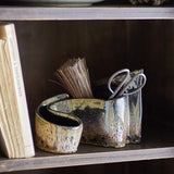 Phil Stoneware Ornament in brown, a curled ceramic piece with open hollows, shown on a shelf holding scissors and dried stems beside a book.