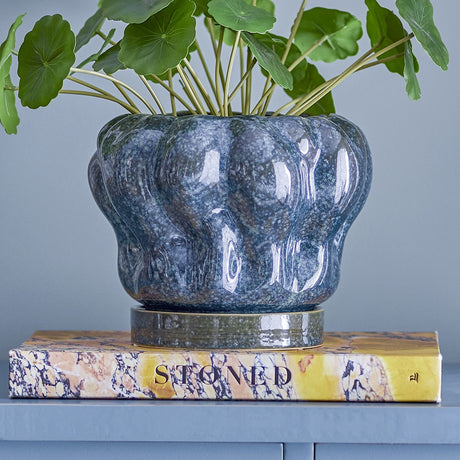 Hollis stoneware plant pot with saucer in glossy blue, featuring a scalloped, lobed rim, shown with greenery on a marbled book in a living room setting.