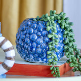 Susannah Stoneware Plant Pot in blue with a textured, pebble-like raised cell pattern, holding a trailing string-of-pearls plant on a styled living room surface.