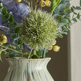Close-up of the Allium Artificial Stem in green, showing its round, spiky faux flower head among eucalyptus foliage and berries in a cream ceramic vase.
