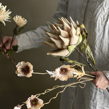 Protea Artificial Stem in beige, held in hand with its layered petal flowerhead and green leaves, shown alongside other faux floral stems against a neutral background.
