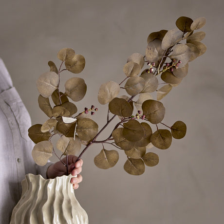 Eucalyptus Artificial Stem 86cm in Green, held in a wavy ceramic vase, showing round coin-shaped leaves and berry clusters on branching stems, in a living room setting.