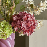 Artificial pink hydrangea stem with a full rounded flowerhead, shown in a floral arrangement alongside green hydrangea blooms and white sprigs in a pink vase.