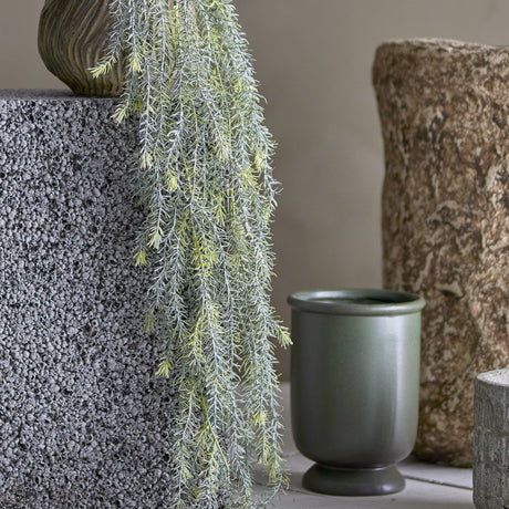 Willow Artificial Plant in green cascading with fine, needle-like trailing foliage from a textured grey vase, styled beside stone decor and a green ceramic pot.