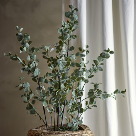 Artificial eucalyptus plant with slender dark stems and airy sprays of green foliage in a textured stone-effect pot, set against a neutral wall and white curtain.