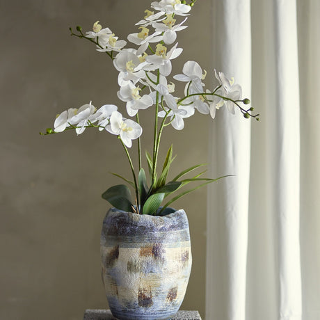 Orchid Artificial Plant in White with arching stems and layered leaves, displayed in a patterned blue-and-cream ceramic pot on a side table near a curtained window.